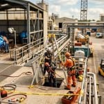 Tilt-shift miniature of a worksite confined-space rescue, with a tripod, winch and standby person, while welding and a forklift continue in the background