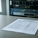 Tilt-shift view of a printed access review list on an office desk with hand-ticked checkboxes, and a rack of powered network switches with lit status indicators out of focus behind it