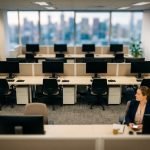 Tilt-shift miniature of an open-plan Australian office with two rows of empty desks and dark monitors, and one person still working at a desk in the foreground.