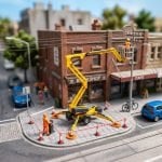 Miniature streetscape with a boom-type elevating work platform on outriggers, its platform raised toward a building facade below overhead powerlines, and a taped exclusion zone on the footpath