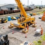 Miniature industrial yard with a mobile crane on outriggers and timber packing inside a taped exclusion zone, and a worker in hi-vis at a table with a lift plan
