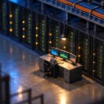 Tilt-shift miniature of a darkened data centre at night, a single figure standing at a plain control desk with three monitors showing abstract graphs, rows of equipment racks with green and amber indicator lights receding into the distance and cable trays overhead