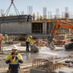 Tilt-shift miniature of a construction site showing a suspended steel load, a slewing excavator, a compressor with pressurised hoses and a concrete saw with water suppression, with a worker in the foreground holding a segmented energy wheel card