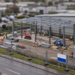 Tilt shift view of a partly built steel frame extension on the side of a warehouse, behind temporary fencing with a blank site sign board facing the road